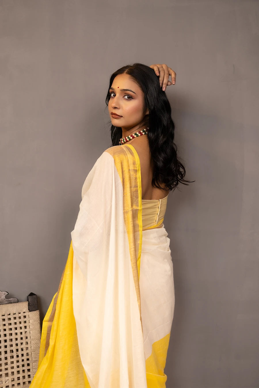 Woman in a yellow and white saree standing next to a chair against a grey background