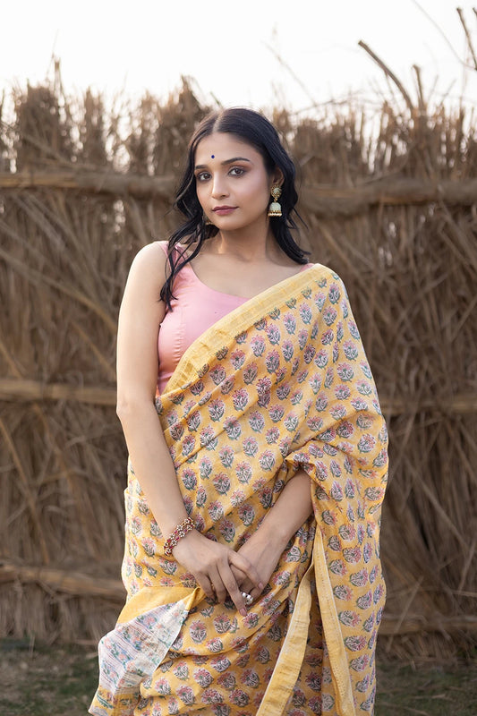 Woman wearing a yellow and pink saree standing in front of a rustic background
