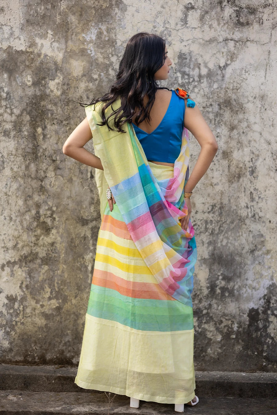 Woman wearing a colorful saree with a blue blouse against a textured wall.