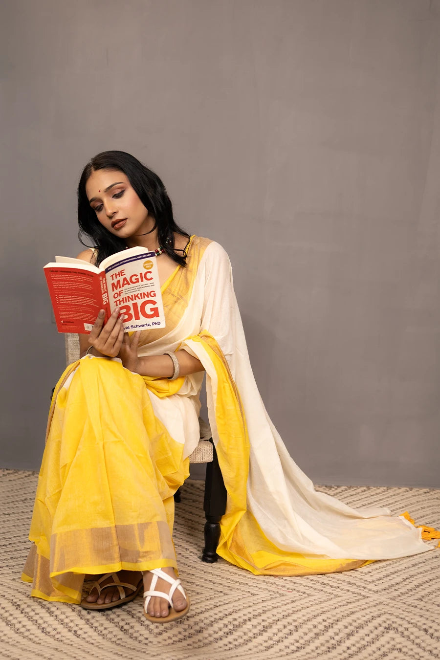 Woman in a yellow and white saree standing next to a chair against a grey background