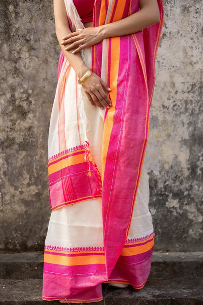 Woman wearing a traditional saree with pink and orange border against a textured wall.