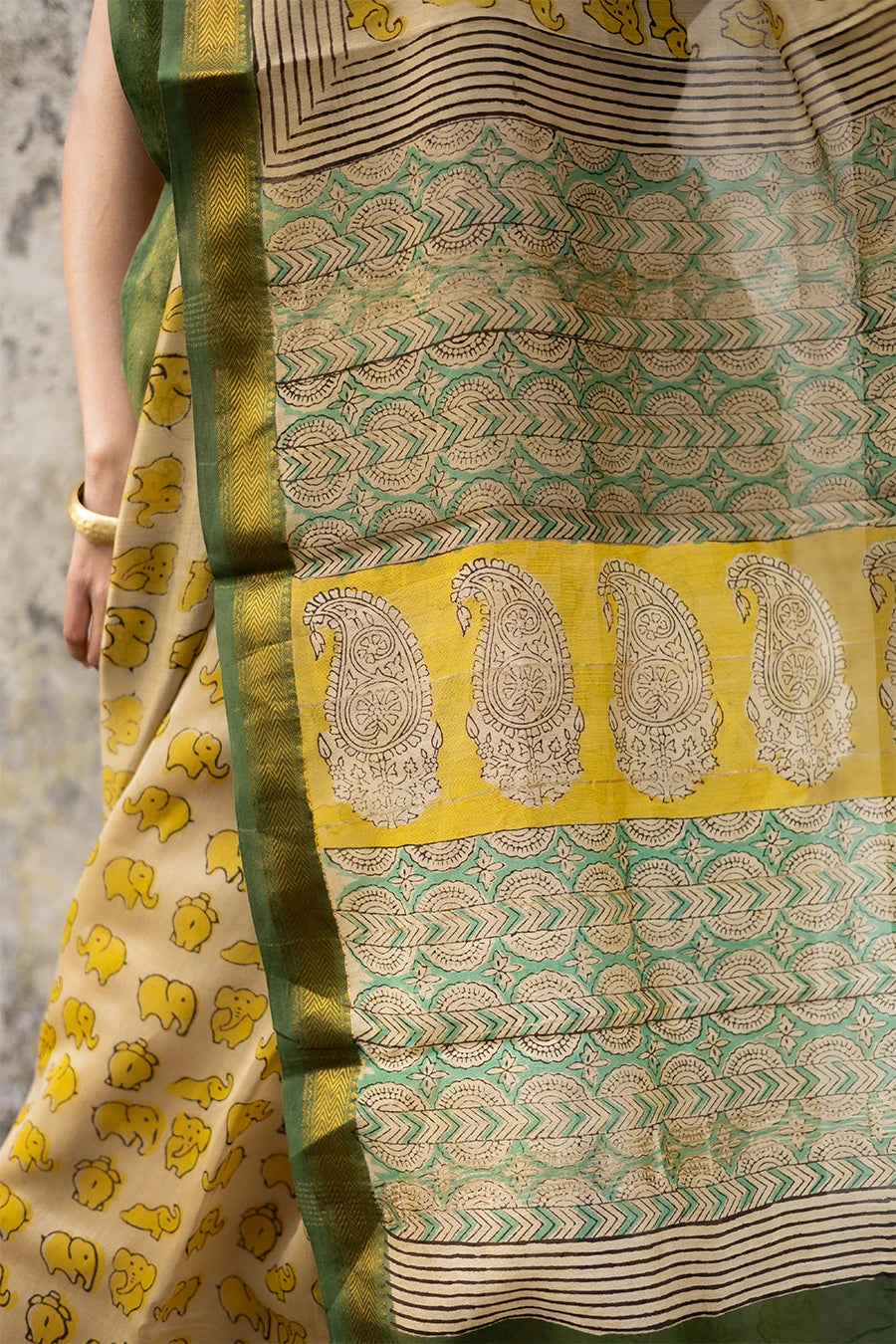 Woman in a yellow and green saree standing against a textured wall.