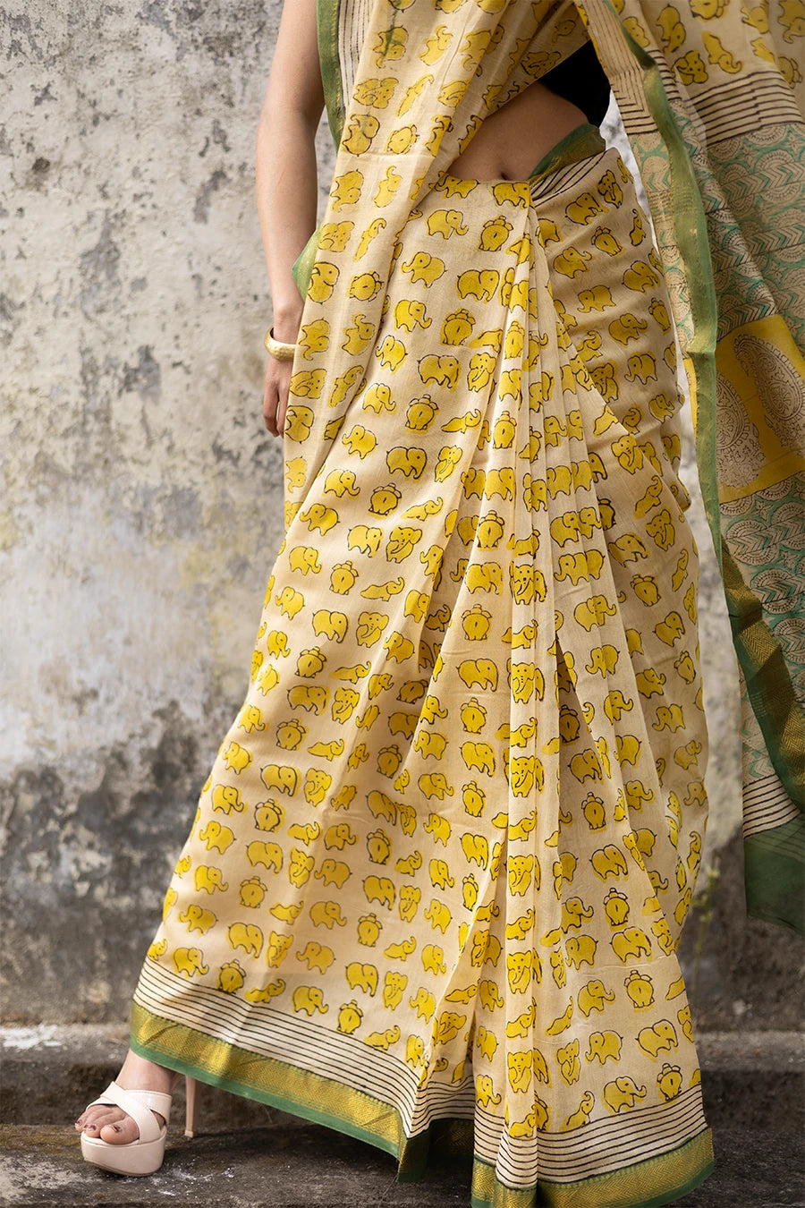 Woman in a yellow and green saree standing against a textured wall.