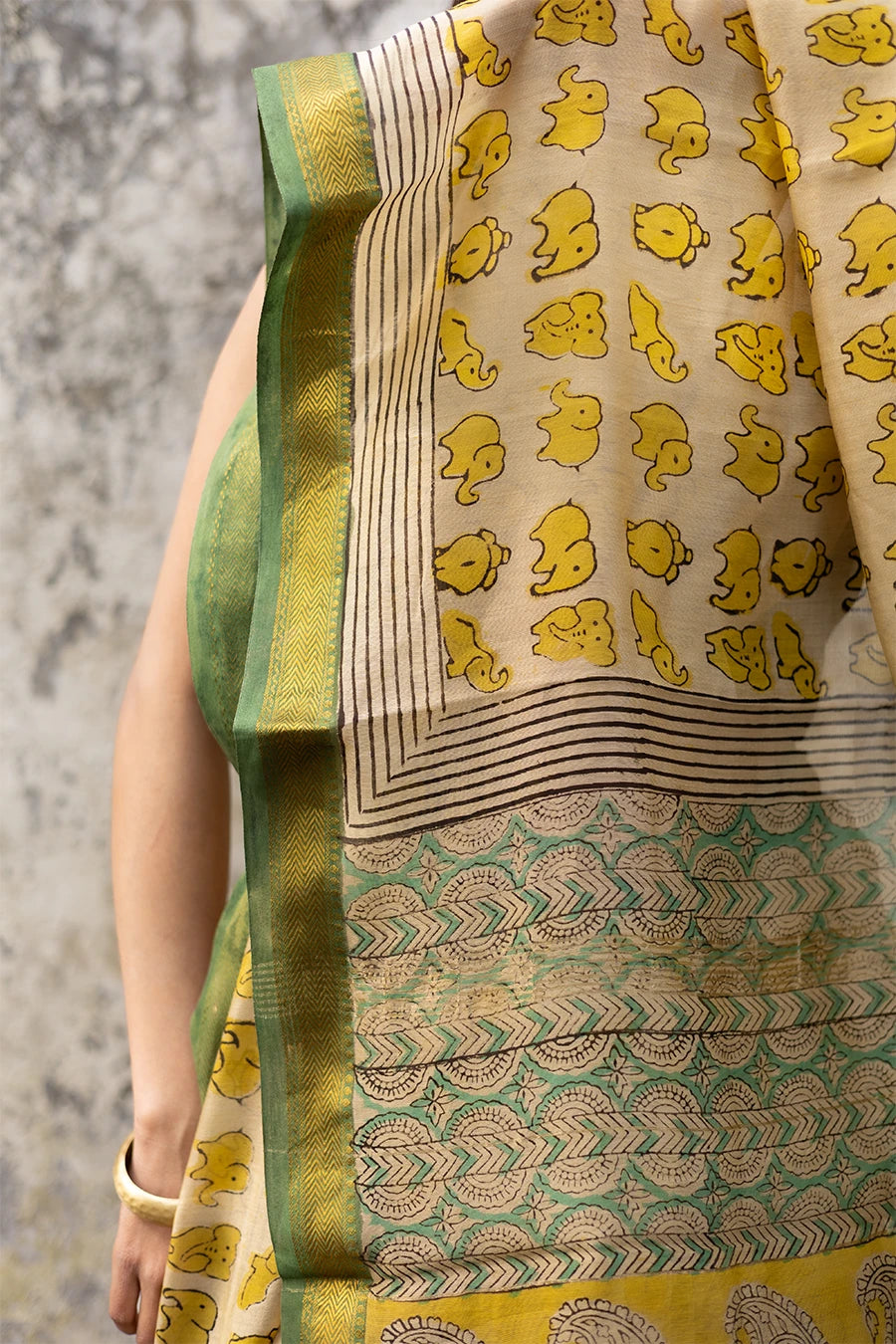 Woman in a yellow and green saree standing against a textured wall.
