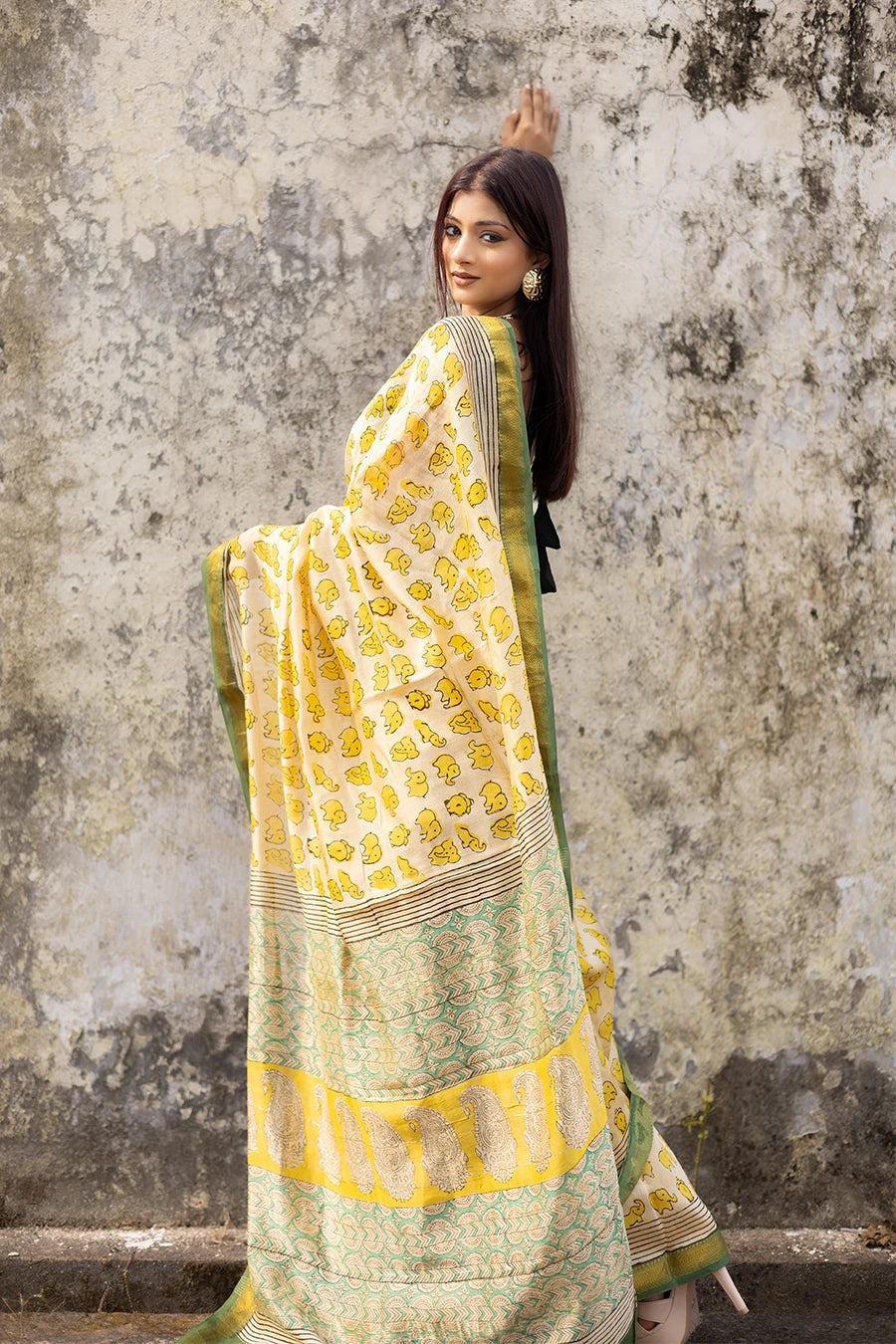 Woman in a yellow and green saree standing against a textured wall.