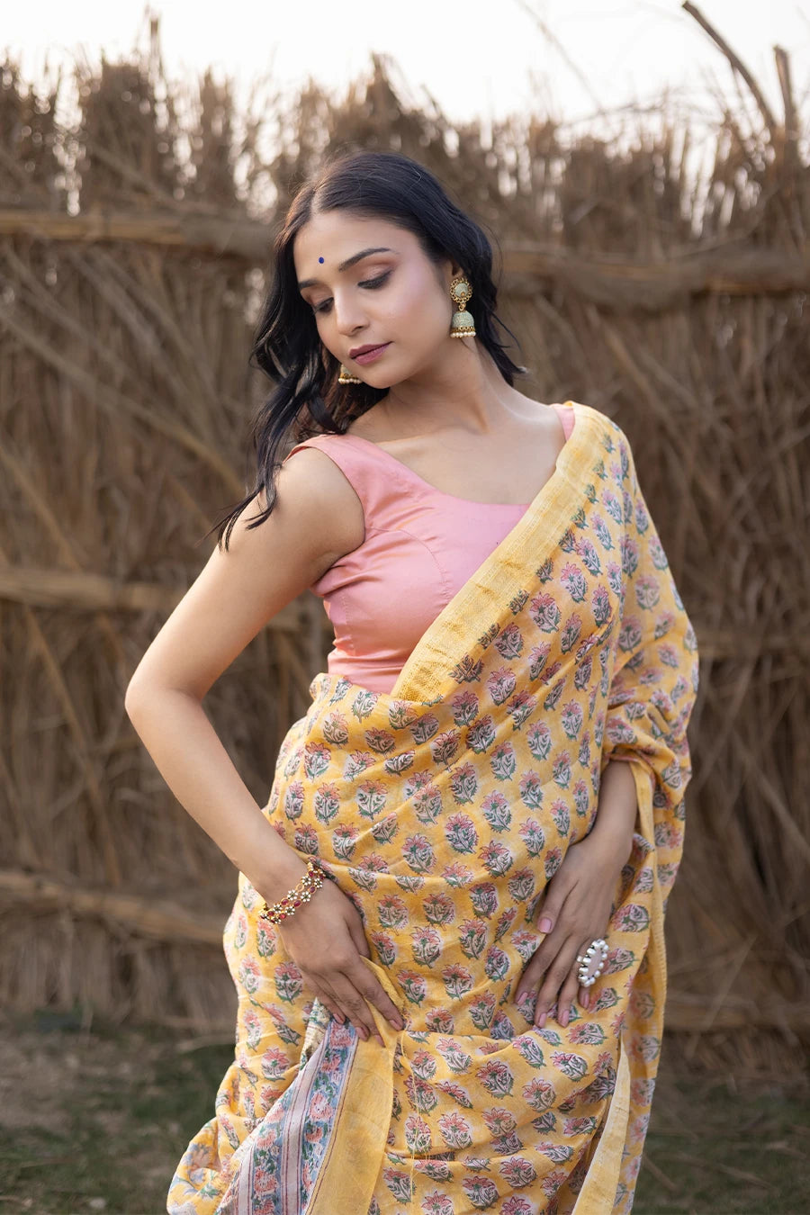 Woman wearing a yellow and pink saree standing in front of a rustic background