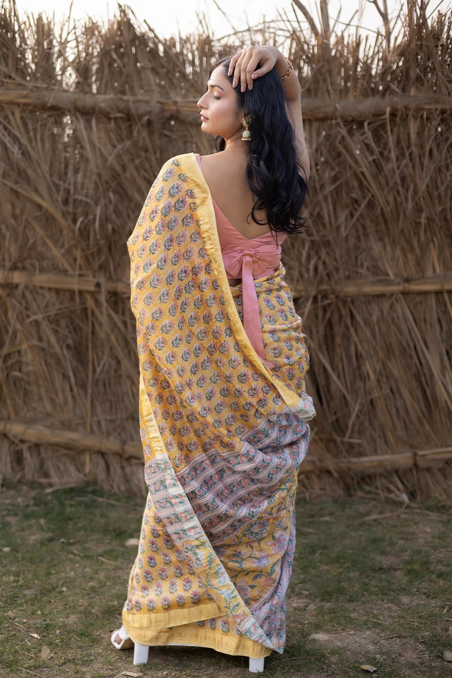 Woman wearing a yellow and pink saree standing in front of a rustic background
