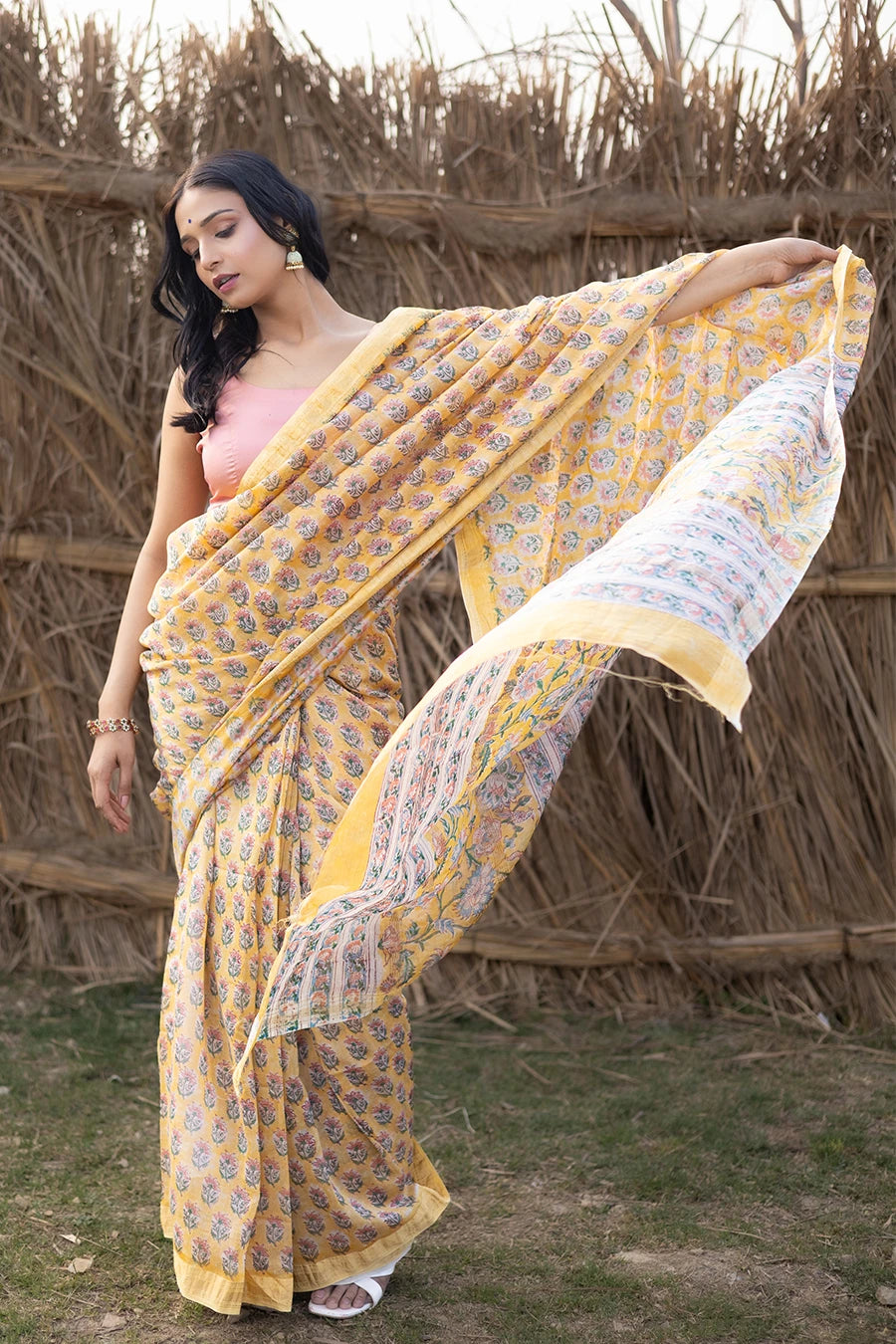 Woman wearing a yellow and pink saree standing in front of a rustic background