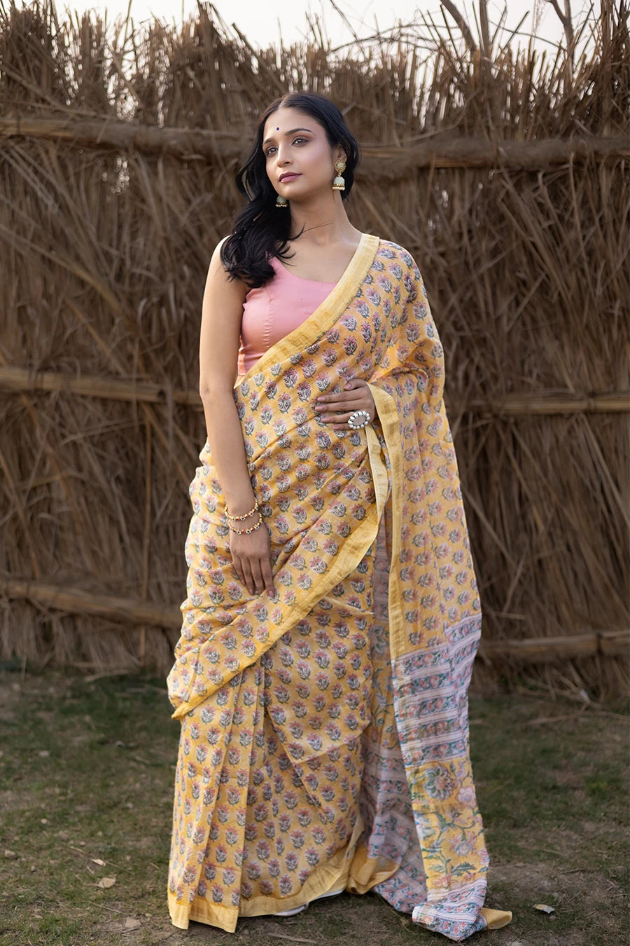 Woman wearing a yellow and pink saree standing in front of a rustic background