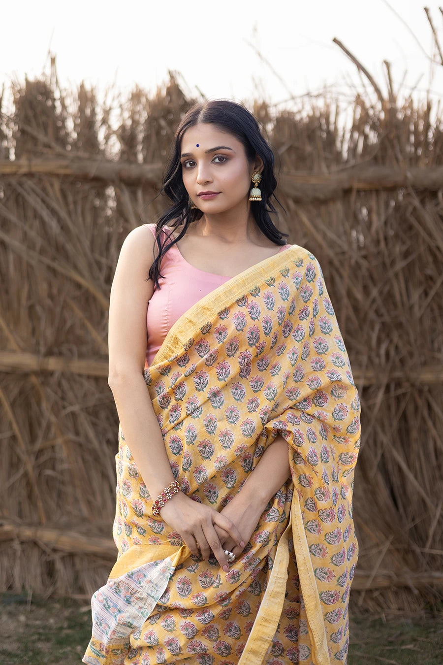 Woman wearing a yellow and pink saree standing in front of a rustic background