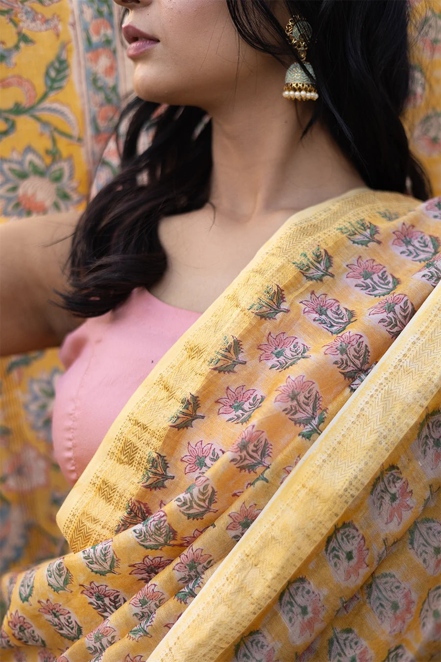 Woman wearing a yellow and pink saree standing in front of a rustic background